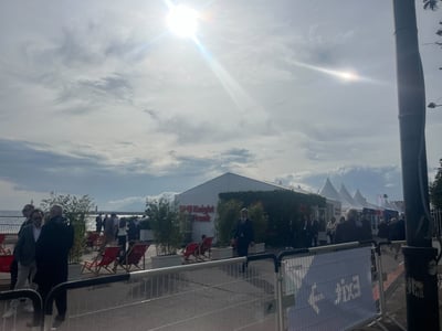 A day at MIPIM March 2026 in Cannes, France. The top two-thirds of the image show a blue, cloudy sky with a sun lens flare. The bottom third features the Knight Frank tent along the promenade, with attendees walking past barriers and several other event tents visible in the background.